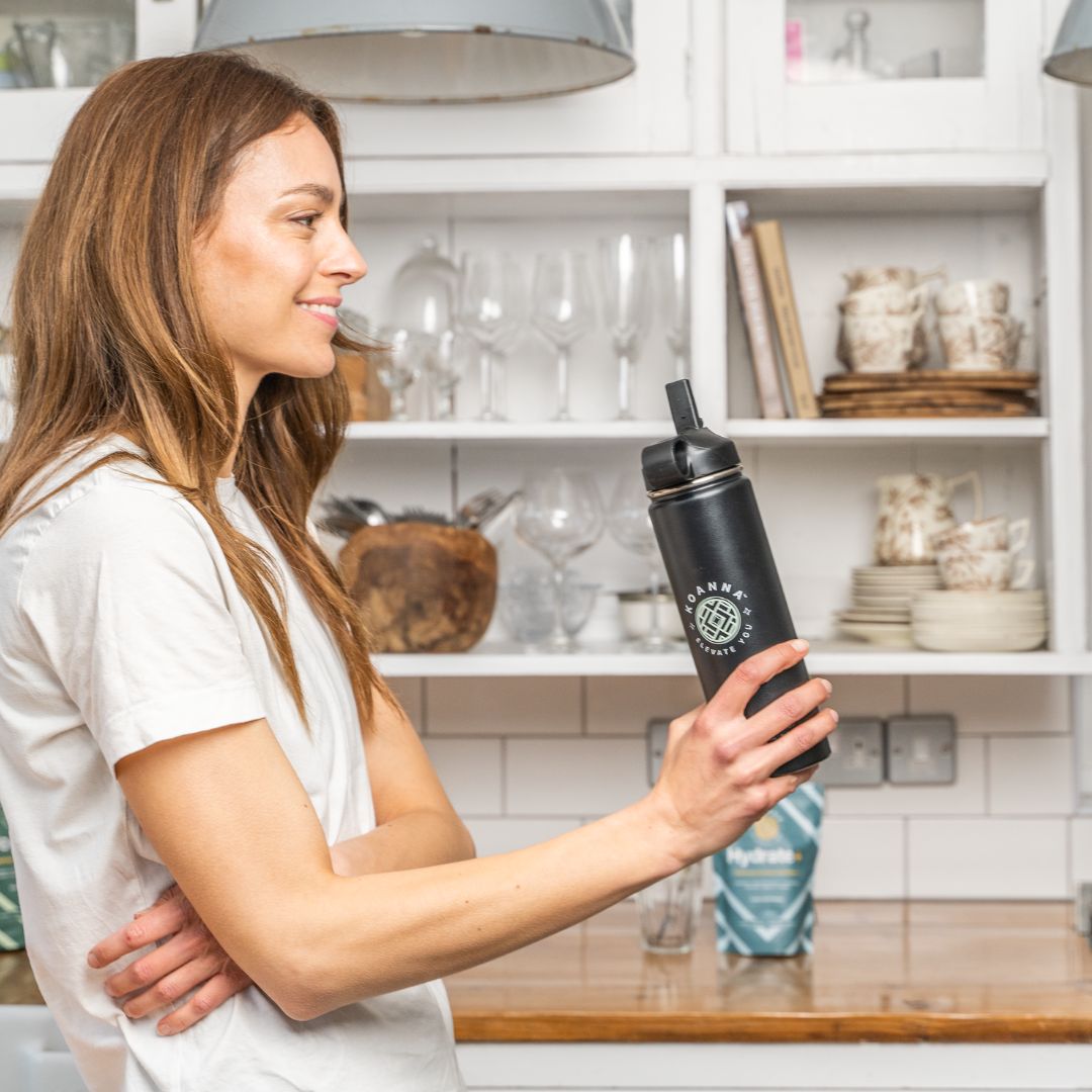 Young woman in her kitchen with black Elevate bottle in hand and Hydrate+ in the background.