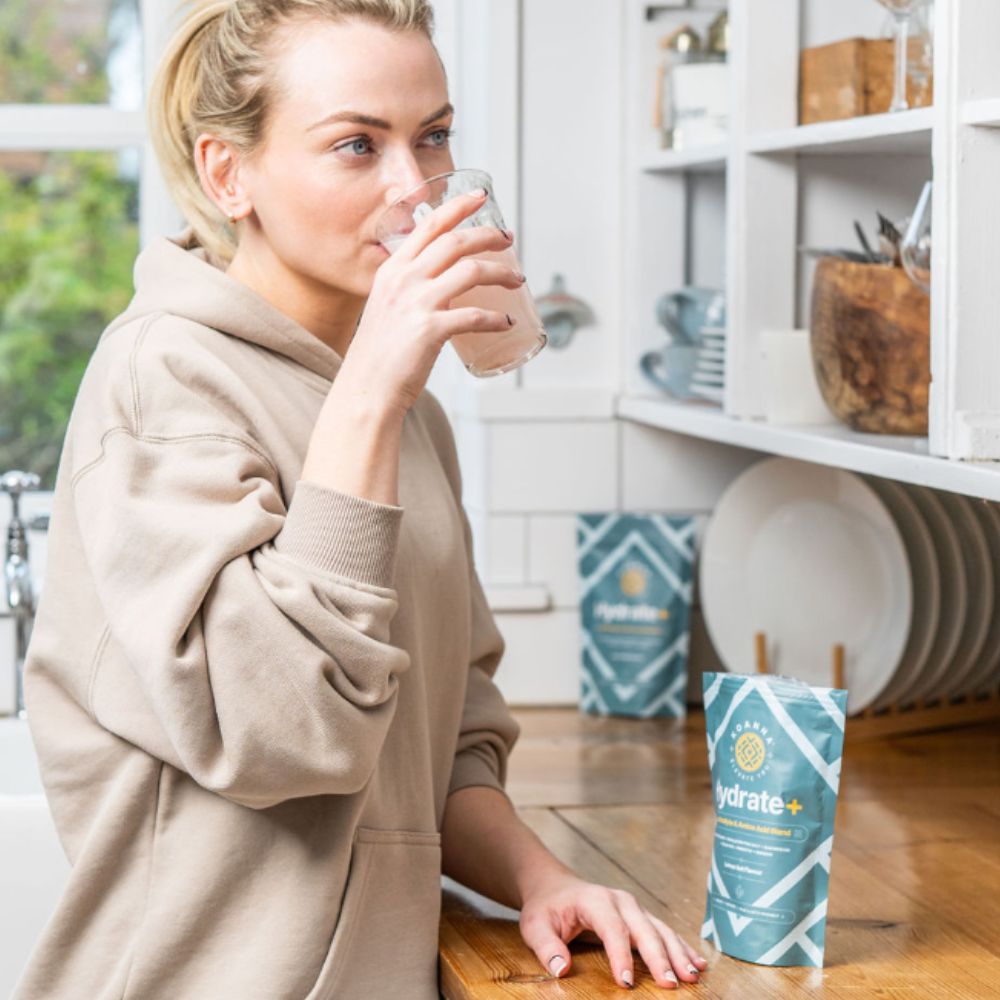 Young blonde woman drinking a glass of Hydrate+ in her kitchen.