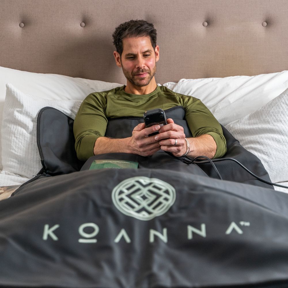 Brown-haired middle-aged man is lying in bed with the black sauna blanket and a book, operating the controller.