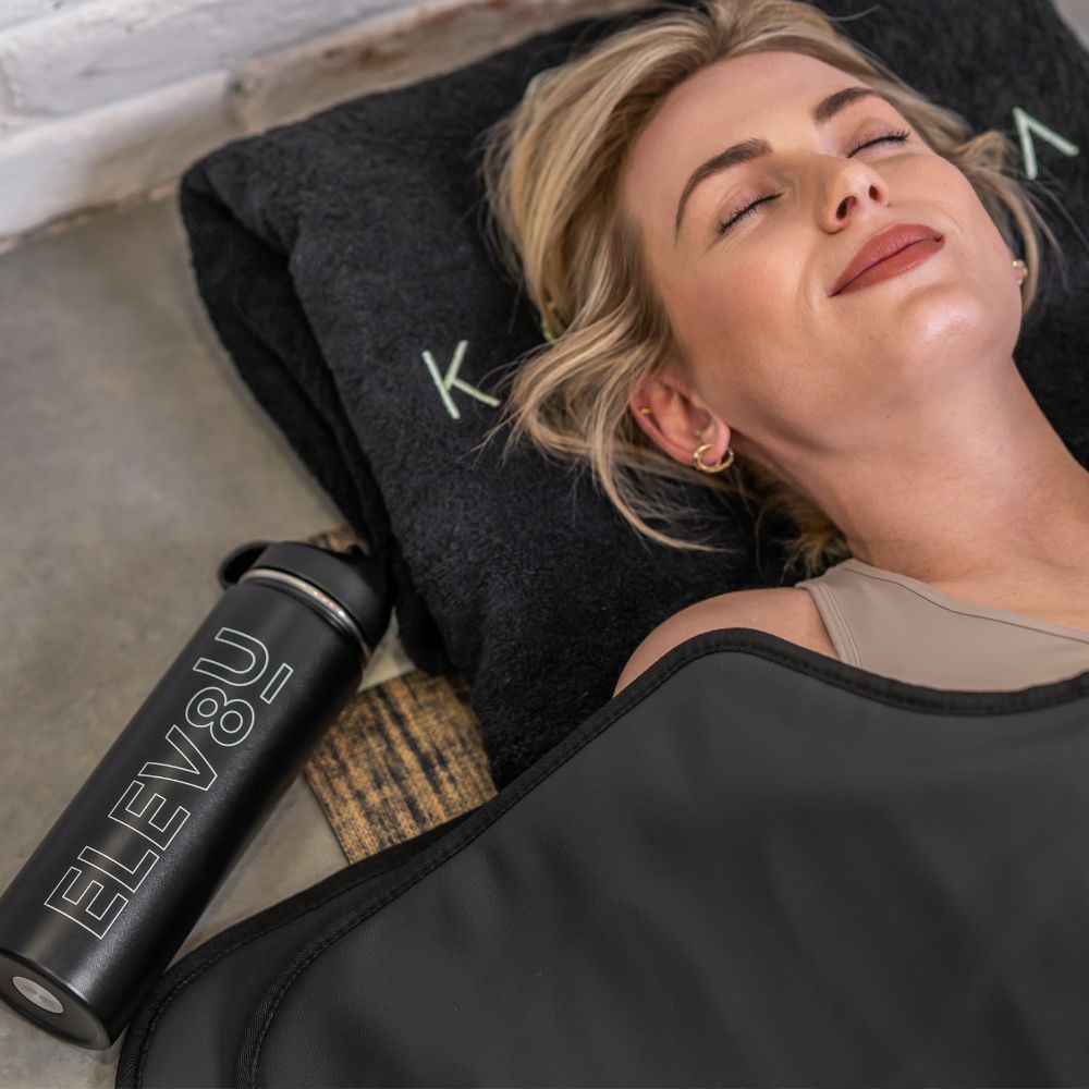 Blonde woman relaxing in her Koanna sauna blanket and next to her the Elevate bottle.